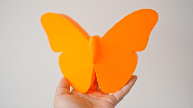 May include: A bright orange butterfly decoration is held in a hand. The butterfly has large, symmetrical wings and a simple, modern design. The vibrant color makes it a striking decorative piece.