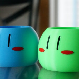 May include: Two round, ceramic planters with a cartoon face design. One planter is blue and the other is green.