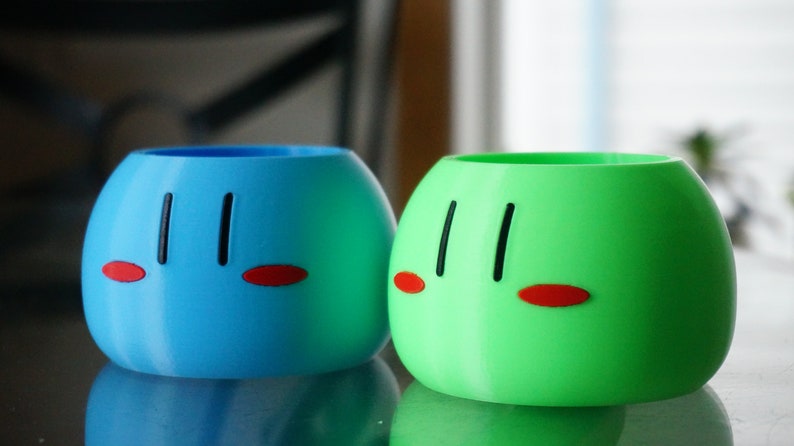 May include: Two round, ceramic planters with a cartoon face design. One planter is blue and the other is green.