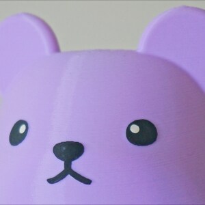 May include: A close-up of a purple bear-shaped toy with black eyes, nose, and mouth.