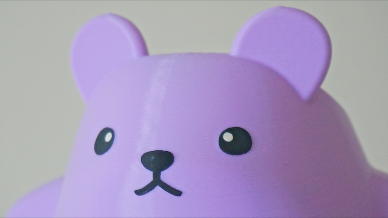 May include: A close-up of a purple bear-shaped toy with black eyes, nose, and mouth.