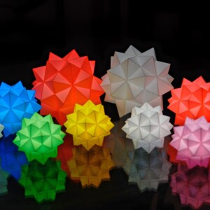 Pode incluir: Uma cole&ccedil;&atilde;o de dez ornamentos de estrelas de papel geom&eacute;tricas em v&aacute;rias cores, incluindo branco, vermelho, azul, verde e amarelo. As estrelas est&atilde;o dispostas em uma fileira, com a maior estrela no centro.