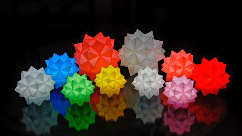 Pode incluir: Uma cole&ccedil;&atilde;o de dez ornamentos de estrelas de papel geom&eacute;tricas em v&aacute;rias cores, incluindo branco, vermelho, azul, verde e amarelo. As estrelas est&atilde;o dispostas em uma fileira, com a maior estrela no centro.
