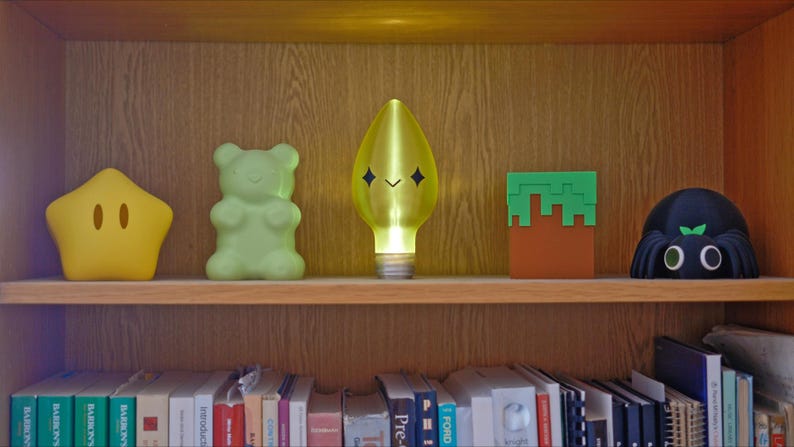 May include: A wooden shelf displays five decorative lights. A yellow star, a green gummy bear, a yellow light bulb with a face, a Minecraft block, and a black spider-shaped light. The shelf is above a row of books.