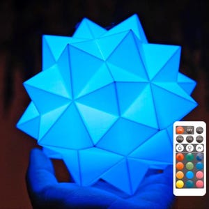 Op de afbeelding: Een blauwe geometrische lamp met een afstandsbediening. De afstandsbediening heeft knoppen voor verschillende kleuren en helderheidsinstellingen.