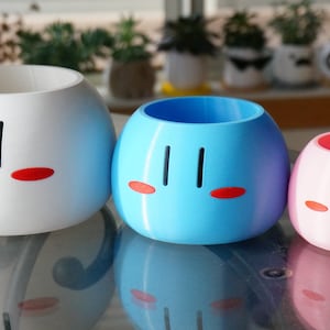 May include: Three ceramic planters shaped like Japanese dango mochi, a sweet rice cake. The planters are white, blue, and pink, and each has a simple face with black eyes and red cheeks.