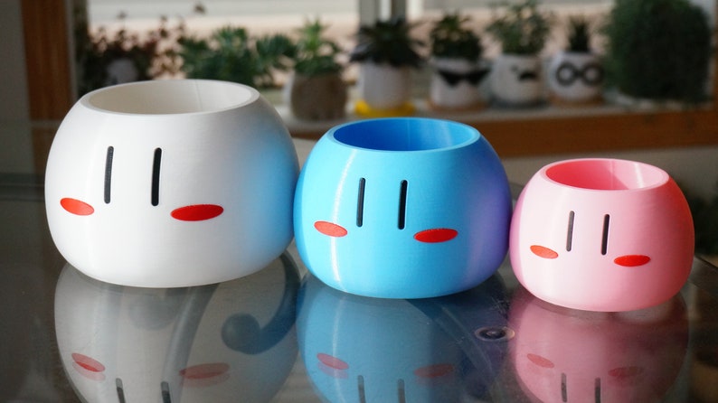 May include: Three ceramic planters shaped like Japanese dango mochi, a sweet rice cake. The planters are white, blue, and pink, and each has a simple face with black eyes and red cheeks.