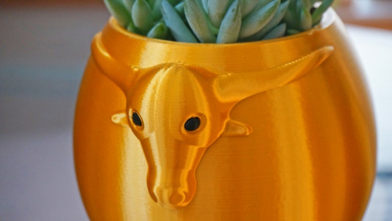 Puede incluir: Una maceta de cer&aacute;mica de color dorado con forma de cabeza de toro con una planta suculenta creciendo en su interior.