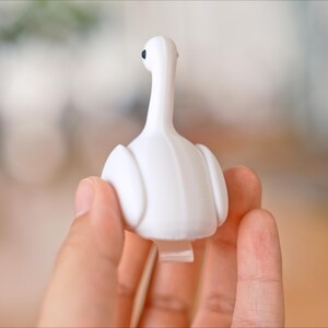May include: A white swan-shaped bottle stopper with a long neck and a small black eye. The stopper is held in a hand, with a blurred background.