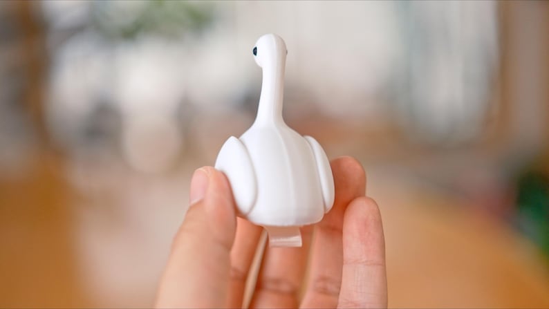 May include: A white swan-shaped bottle stopper with a long neck and a small black eye. The stopper is held in a hand, with a blurred background.