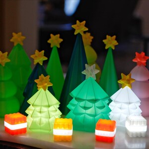 May include: A collection of illuminated Christmas tree decorations in various shades of green, white, and pink. Each tree is topped with a yellow star. Small, glowing, square-shaped lights in red, white, and yellow are also present, creating a festive holiday display.