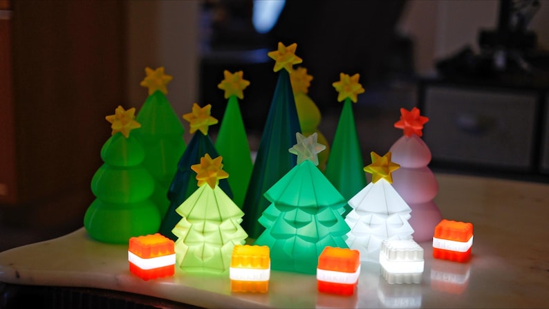 May include: A collection of illuminated Christmas tree decorations in various shades of green, white, and pink. Each tree is topped with a yellow star. Small, glowing, square-shaped lights in red, white, and yellow are also present, creating a festive holiday display.