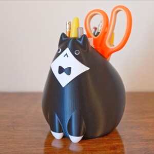May include: A black cat-shaped desk organizer with a white tuxedo and bow tie. The organizer holds orange scissors, pencils, and pens. The cat has white feet and is sitting on a wooden surface.