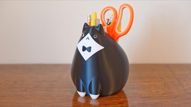 May include: A black cat-shaped desk organizer with a white tuxedo and bow tie. The organizer holds orange scissors, pencils, and pens. The cat has white feet and is sitting on a wooden surface.