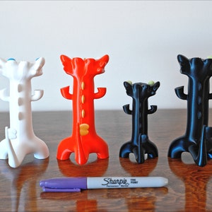 May include: Four colorful, stylized dragon-shaped pen holders in white, red, and black. A purple Sharpie pen is in front of the holders for size comparison. The holders have a unique, decorative design.