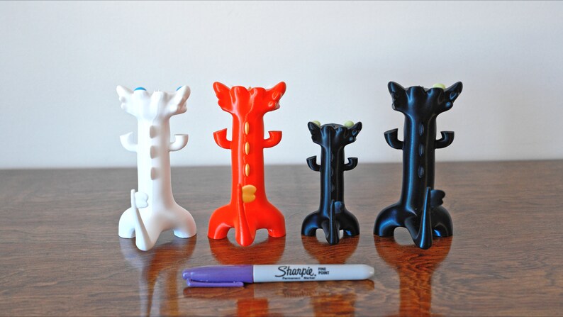 May include: Four colorful, stylized dragon-shaped pen holders in white, red, and black. A purple Sharpie pen is in front of the holders for size comparison. The holders have a unique, decorative design.
