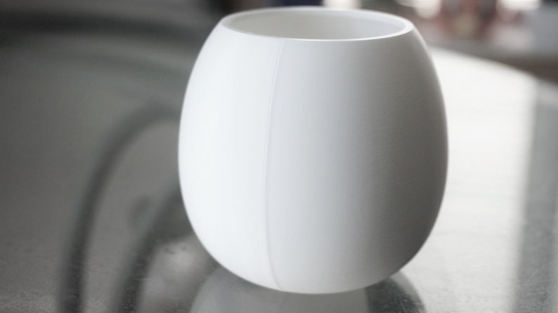 May include: A white ceramic vase with a smooth, rounded shape. The vase has a subtle seam down the side.