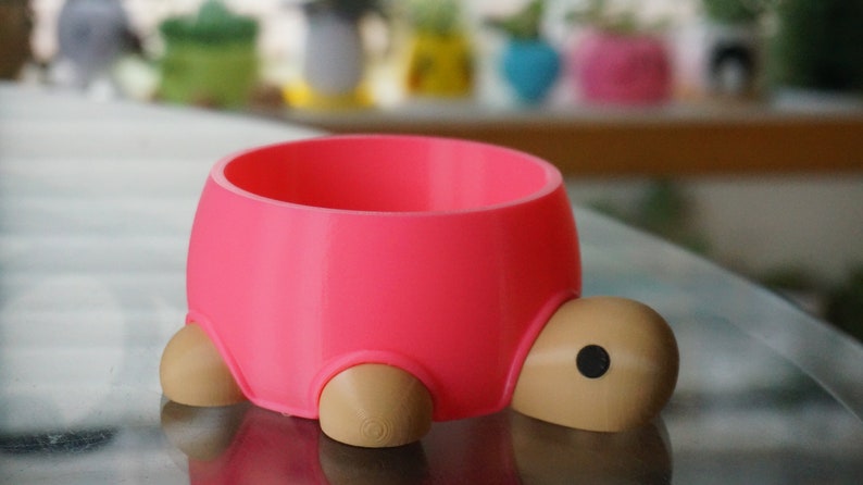 May include: A pink turtle-shaped planter with brown legs and a black eye. The planter is made of plastic and is designed to hold small plants.