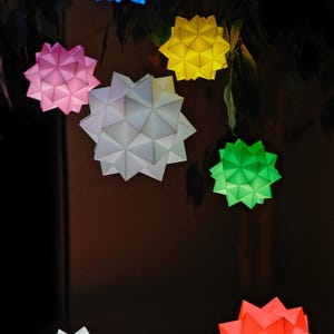 Pode incluir: Uma cole&ccedil;&atilde;o de oito estrelas de papel origami em v&aacute;rias cores, incluindo azul, rosa, amarelo, branco, verde e vermelho. As estrelas est&atilde;o penduradas em um galho de &aacute;rvore e s&atilde;o iluminadas por dentro.