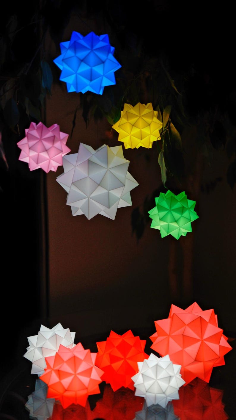 Pode incluir: Uma cole&ccedil;&atilde;o de oito estrelas de papel origami em v&aacute;rias cores, incluindo azul, rosa, amarelo, branco, verde e vermelho. As estrelas est&atilde;o penduradas em um galho de &aacute;rvore e s&atilde;o iluminadas por dentro.