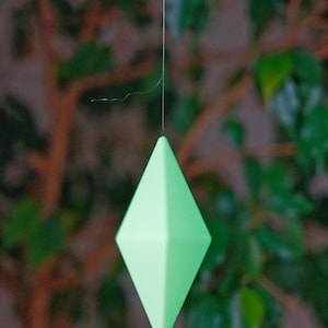 May include: A green diamond-shaped ornament hanging from a string against a blurred background of green leaves.