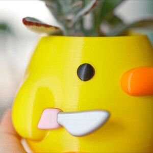 May include: A yellow, duck-shaped planter with a black eye, orange cheek, and a pink and silver beak. The planter holds a small succulent plant. The design is whimsical and playful, suitable for home decor.
