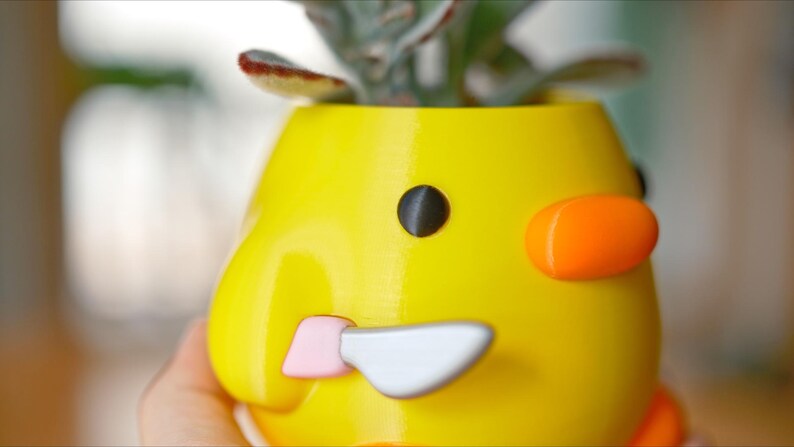 May include: A yellow, duck-shaped planter with a black eye, orange cheek, and a pink and silver beak. The planter holds a small succulent plant. The design is whimsical and playful, suitable for home decor.