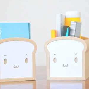 May include: Two beige bread-shaped desk organizers with white faces. One organizer has a blue notebook inside and the other has a stack of papers, a blue pen, a yellow highlighter, and a silver stapler.