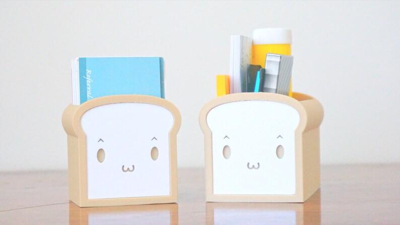 May include: Two beige bread-shaped desk organizers with white faces. One organizer has a blue notebook inside and the other has a stack of papers, a blue pen, a yellow highlighter, and a silver stapler.