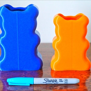 Puede incluir: Dos jarrones con forma de oso impresos en 3D, uno azul y otro naranja, est&aacute;n sobre una superficie de madera. Un marcador azul est&aacute; delante de los jarrones.