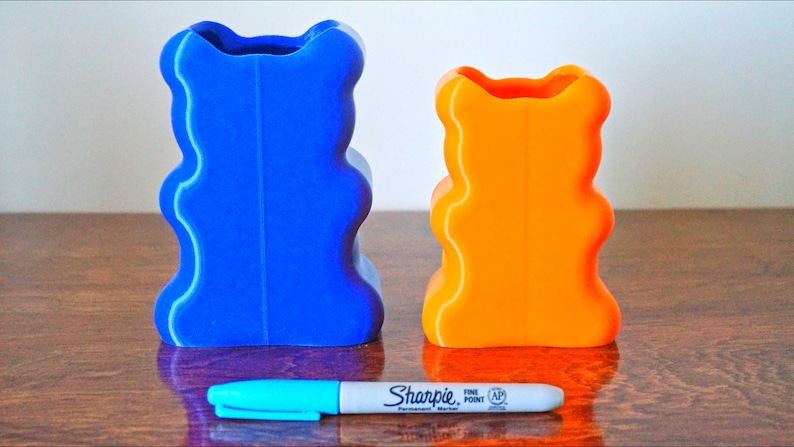 Puede incluir: Dos jarrones con forma de oso impresos en 3D, uno azul y otro naranja, est&aacute;n sobre una superficie de madera. Un marcador azul est&aacute; delante de los jarrones.