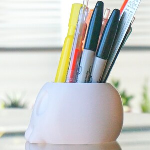 May include: White ceramic skull-shaped pencil holder with a variety of pens and markers inserted. The holder is sitting on a glass surface.