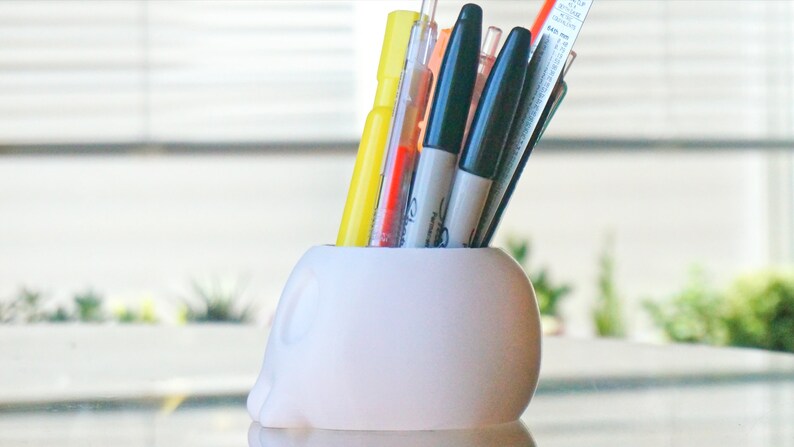 May include: White ceramic skull-shaped pencil holder with a variety of pens and markers inserted. The holder is sitting on a glass surface.