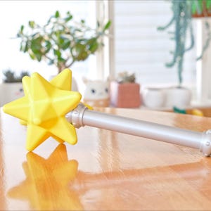 May include: A yellow star-shaped wand with a silver handle rests on a wooden table. The star has a faceted design, and the handle has decorative silver accents. The wand is a toy or prop.