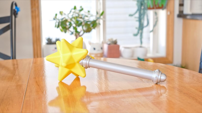 May include: A yellow star-shaped wand with a silver handle rests on a wooden table. The star has a faceted design, and the handle has decorative silver accents. The wand is a toy or prop.