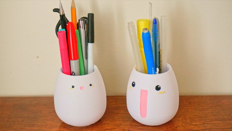 May include: Two white ceramic egg-shaped pencil holders with cute faces. One holder has a pink tongue and the other has a pink nose. Both holders are filled with various pens, pencils, and markers.