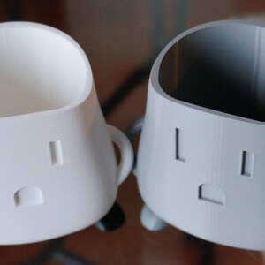 Op de afbeelding: Twee 3D-geprinte keramische bekers met een witte en grijze afwerking. De bekers hebben een uniek ontwerp met een gezicht met twee gaten en een mond. De bekers zijn perfect om kleine items zoals pennen, potloden of andere bureauaccessoires in te bewaren.