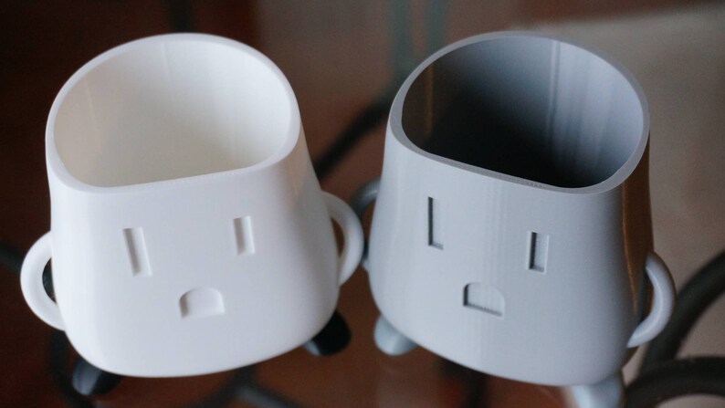 Op de afbeelding: Twee 3D-geprinte keramische bekers met een witte en grijze afwerking. De bekers hebben een uniek ontwerp met een gezicht met twee gaten en een mond. De bekers zijn perfect om kleine items zoals pennen, potloden of andere bureauaccessoires in te bewaren.