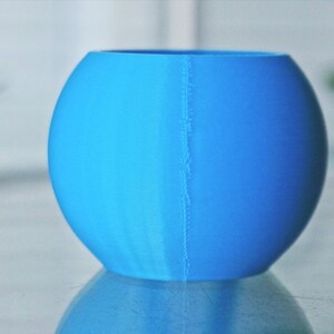 May include: A bright blue, round, ceramic planter with a seam down the side. The planter is sitting on a reflective surface.