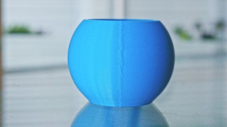 May include: A bright blue, round, ceramic planter with a seam down the side. The planter is sitting on a reflective surface.