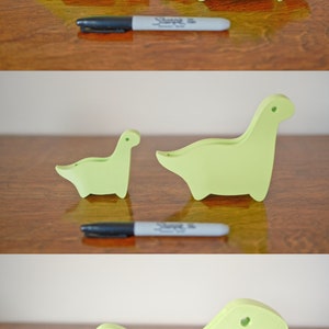 May include: Three green dinosaur-shaped doorstops of varying sizes. The largest doorstop is a stegosaurus with plates on its back. The middle doorstop is a brachiosaurus with a long neck. The smallest doorstop is a tyrannosaurus rex with a short neck and small arms.