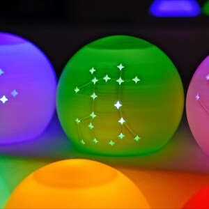 May include: Glowing, spherical decorative lights in various colors, including purple, green, and pink. Each light features a constellation design, emitting a soft, ambient glow. The lights are arranged in a row, creating a colorful and celestial display.