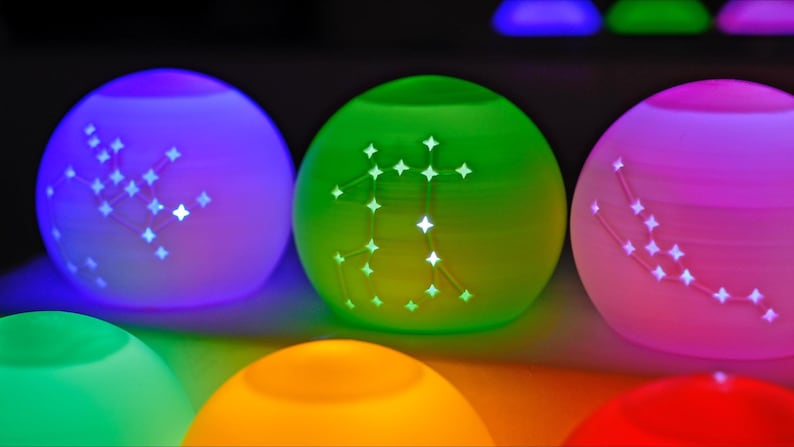 May include: Glowing, spherical decorative lights in various colors, including purple, green, and pink. Each light features a constellation design, emitting a soft, ambient glow. The lights are arranged in a row, creating a colorful and celestial display.