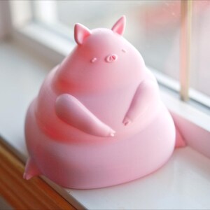 May include: A pink, chubby pig figurine with a rounded body and small ears. The pig is sitting on a white surface.