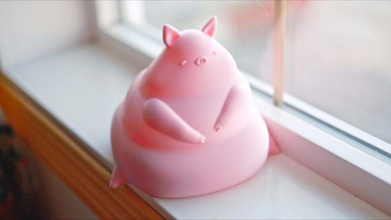 May include: A pink, chubby pig figurine with a rounded body and small ears. The pig is sitting on a white surface.