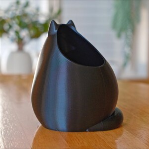 May include: A black cat-shaped planter with a smooth, matte finish. The planter has a large opening for plants and a curled tail. The cat planter is sitting on a wooden table.