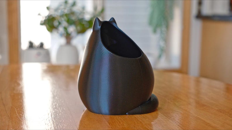 May include: A black cat-shaped planter with a smooth, matte finish. The planter has a large opening for plants and a curled tail. The cat planter is sitting on a wooden table.