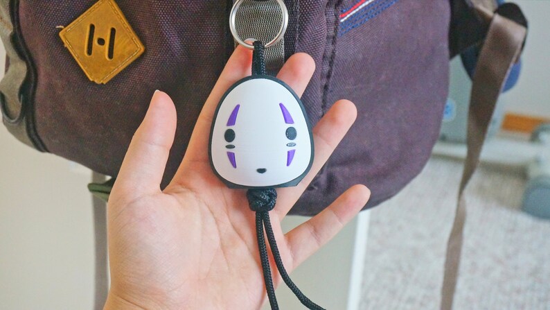 May include: A white and black keychain featuring a character face with purple accents. The keychain is attached to a black cord and a silver ring. The background includes a purple backpack.