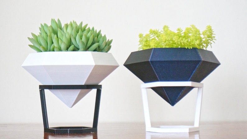 May include: Two diamond-shaped planters, one white and one black, with succulent plants inside. Each planter sits on a geometric stand.