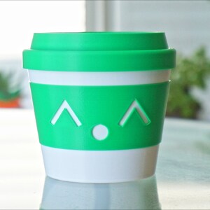 May include: A green and white reusable coffee cup with a cute face. The cup has a white band around the middle and a green lid.